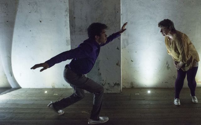 A photo still of the performance Empty Theatre Dream by Corali Dance Company 2014. The photo is taken in Dilston Grove (Café Gallery Projects) and features Jackie Ryan  and Housni Hassan (DJ) who dance in front of the stage and create shadows against the white wall, their costumes are burgundy and gold.